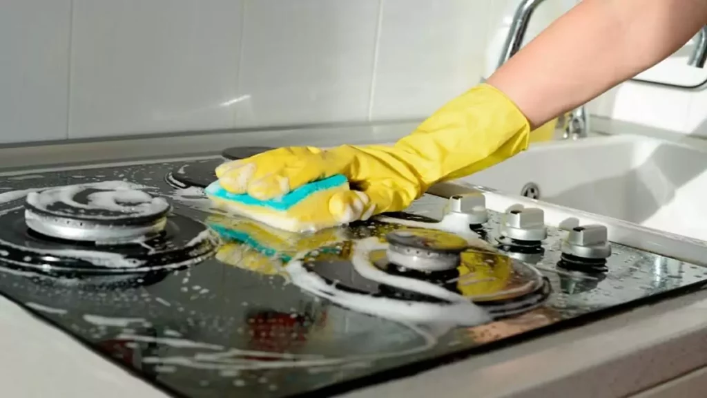 Abertura do post sobre como limpar fogão de vidro: Mulher limpando fogão com luva amarela e esponja verde